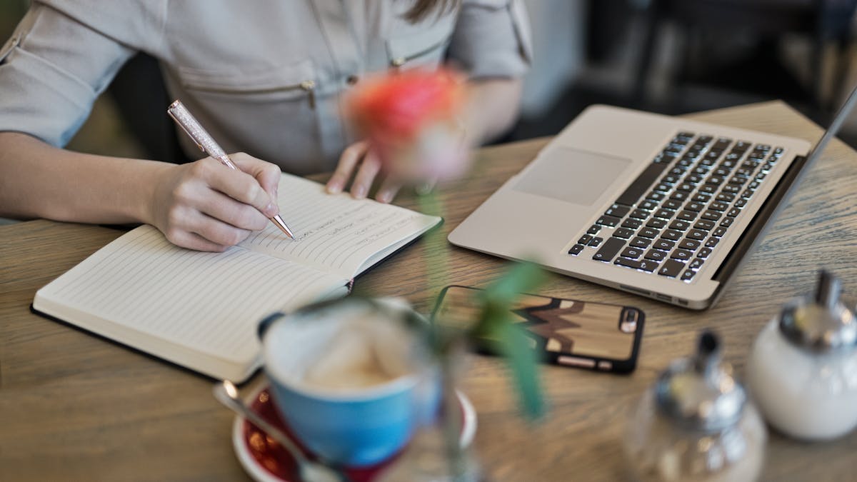 A person writing in a notebook beside a laptop